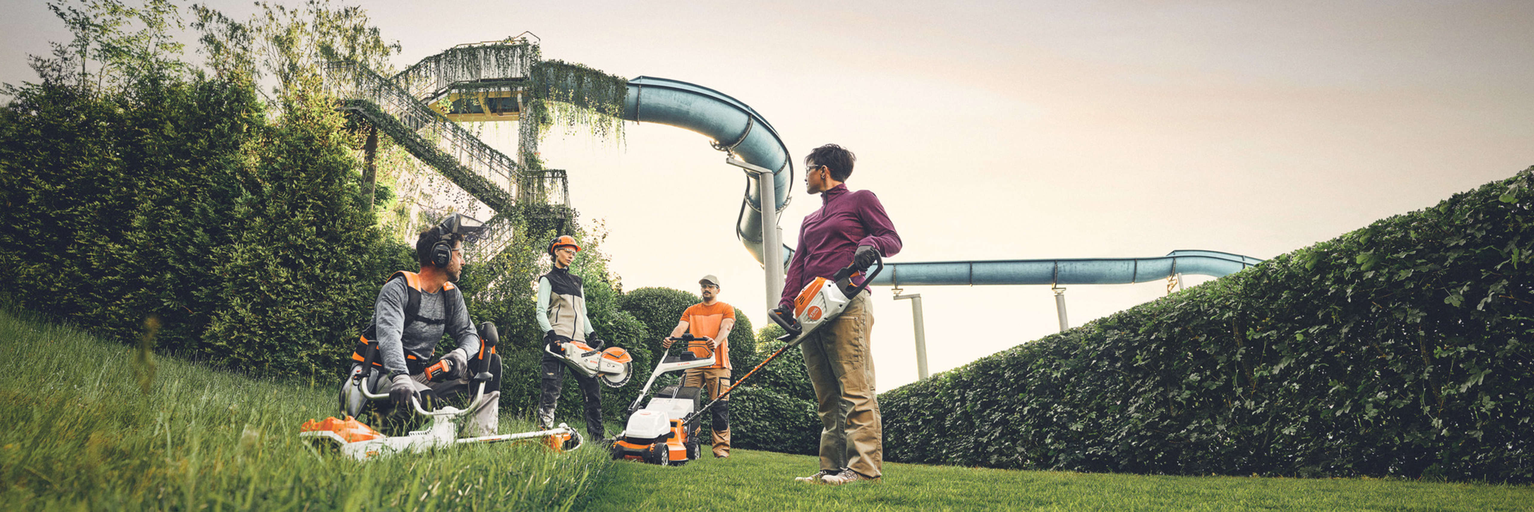 Grupa ljudi radi sa STIHL baštenskim alatima na zelenoj površini sa velikom živom ogradom i vodenim toboganima u pozadini. Opremljeni su trimerima, kosilicama i zaštitnom opremom.